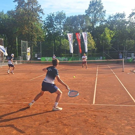 Ein Tennismatch auf einem Sandplatz mit Spielern im Hintergrund. Ein Spieler ist kurz davor, den Ball zu schlagen.