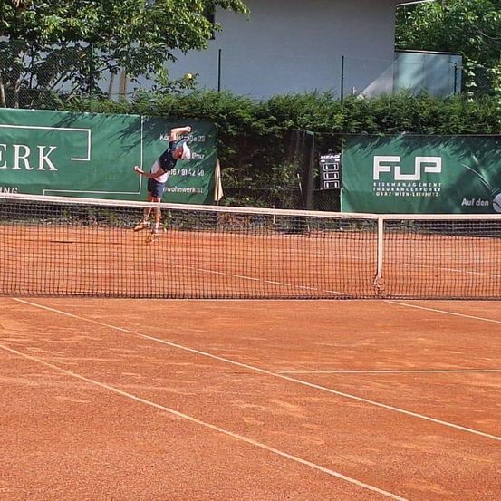 Ein Tennisspieler schwingt nach einem Tennisball auf einem Außentennisplatz. Der Platz ist von einem Zaun umgeben, an dem Werbung angebracht ist.