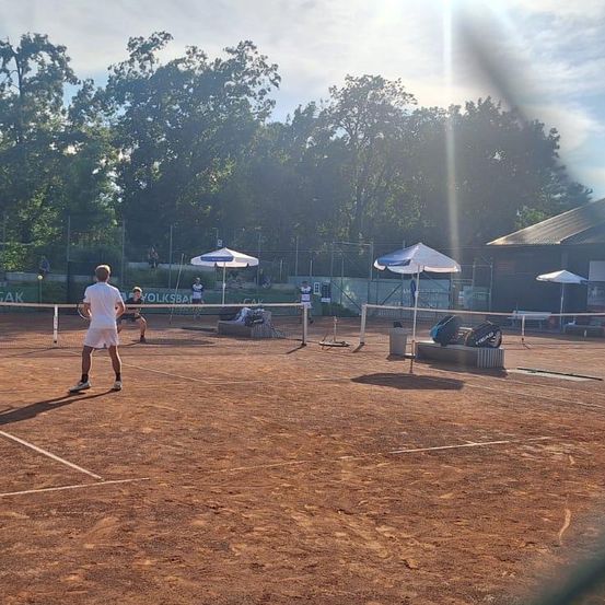 Ein Tennisspieler steht auf einem Platz mit einem Netz, während sich eine Person hinter dem Netz duckt. Zwei Schirme sind vorhanden, mit Tennistaschen auf einer nahen Bank.