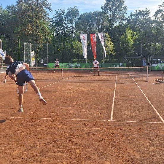 Tennisspieler auf einem Außenplatz, einer in der Luft, ein anderer mit einem Tennisschläger, und zwei Fahnen im Hintergrund.