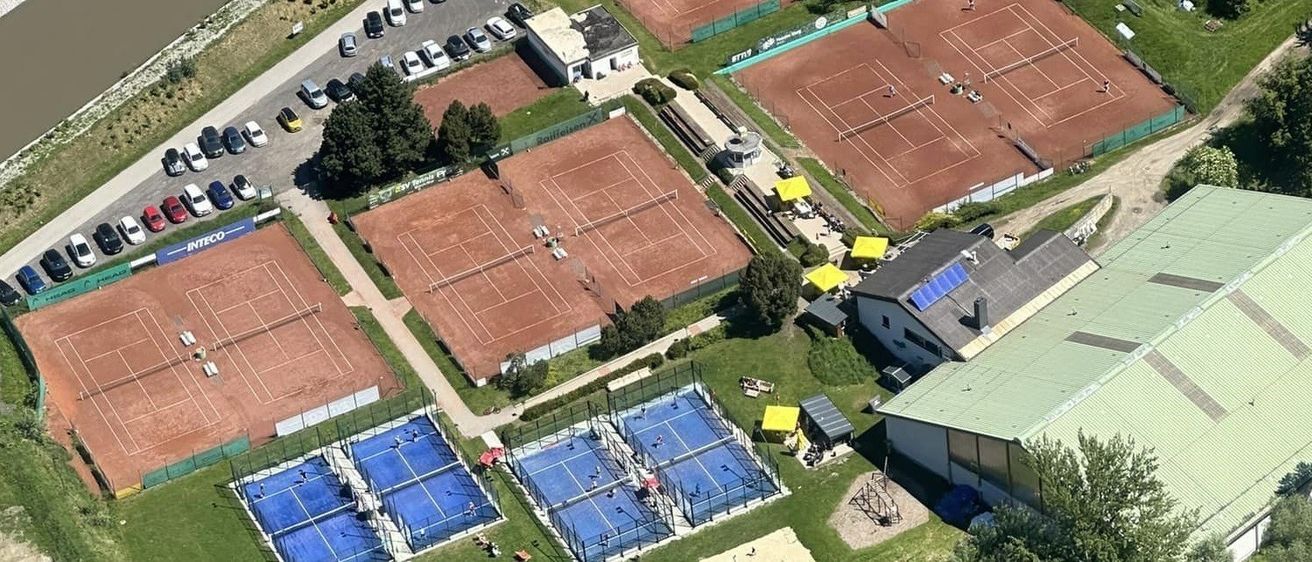 Luftaufnahme einer Tennisanlage mit mehreren Plätzen, darunter ein Hartplatz, umgeben von Bäumen und einem Parkplatz mit mehreren Autos.