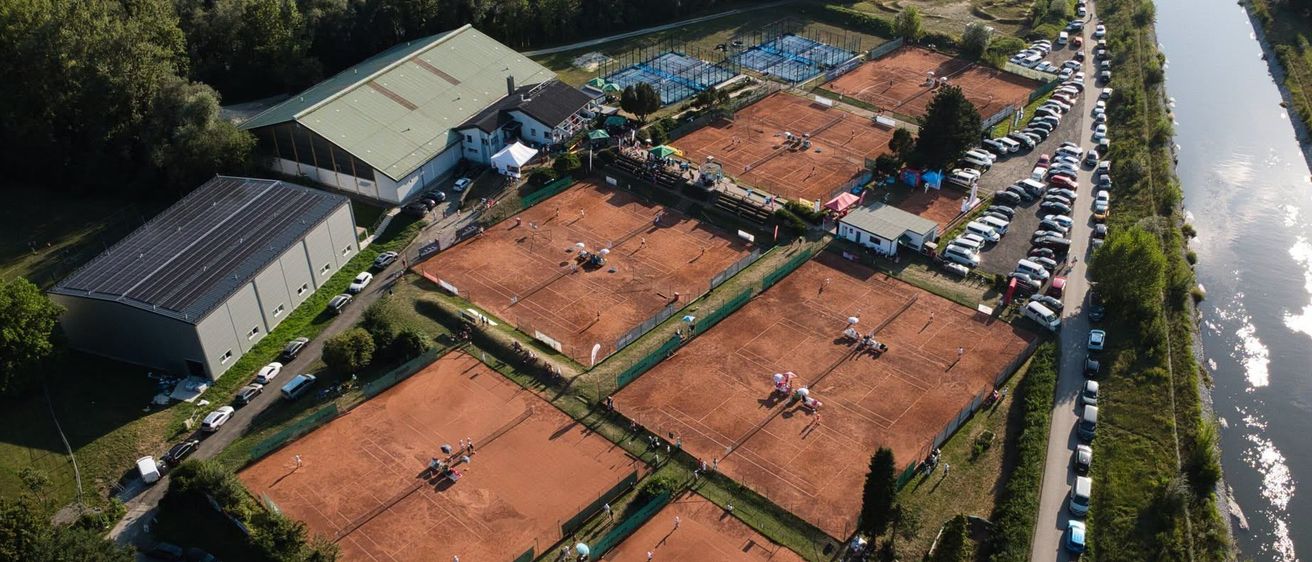 Eine Luftaufnahme eines Tenniskomplexes mit mehreren Plätzen, Zuschauern und einem Parkplatz. Gebäude und Bäume umgeben die Plätze.