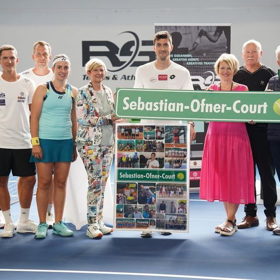 Eine Gruppe von Menschen posiert für ein Foto auf einem Tennisplatz mit einem Schild, das Sebastian-Ofner-Court steht. Sie alle lächeln und scheinen zu feiern.