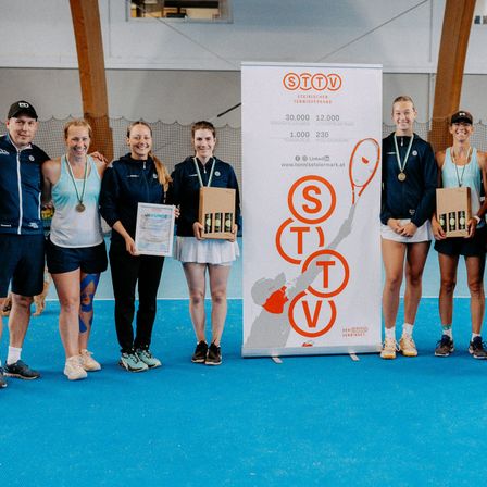 Sechs Personen stehen auf einem Tennisplatz und halten Auszeichnungen und Schachteln. Sie tragen Medaillen und halten Urkunden. Ein großer Banner hinter ihnen hat die Buchstaben STTV.