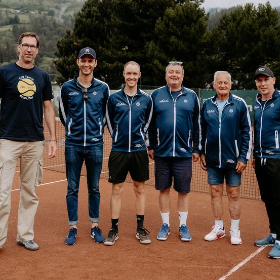 Fünf Männer in blauen Jacken und Turnschuhen stehen auf einem Tennisplatz. Sie scheinen für ein Foto zu posieren.