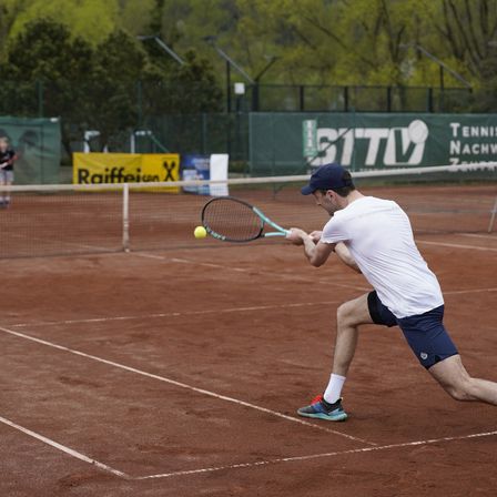 Ein Mann in einem weißen T-Shirt und blauen Shorts spielt Tennis und schlägt einen gelben Ball mit seinem Schläger auf einem Sandplatz. Der Platz ist von einem grünen Zaun mit einem Sponsorenbanner umgeben.