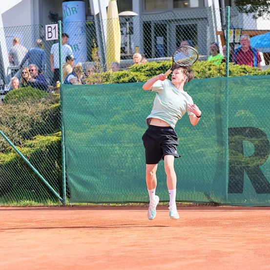 Ein Tennisspieler springt mit einem Schläger in der Luft auf einem Tennisplatz. Hinter ihm ist ein grüner Zaun und ein großer grüner Buchstabe R zu sehen. Zuschauer beobachten von der anderen Seite des Zauns.