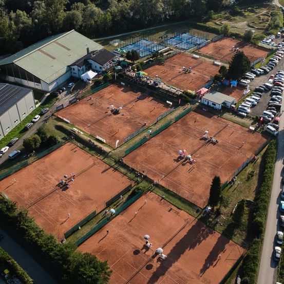 Luftaufnahme eines Tenniskomplexes mit mehreren Plätzen, Zuschauern und geparkten Autos. Gebäude und Bäume sind im Hintergrund zu sehen.