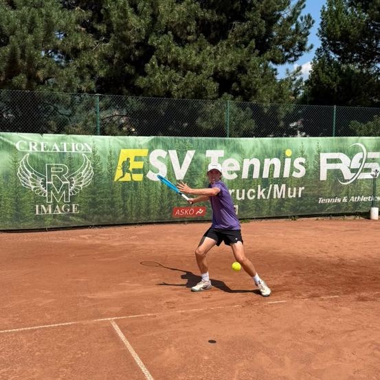 Ein Mann in einem lila Shirt und schwarzen Shorts schwingt einen Tennisschläger auf einem Sandplatz mit einem grünen Banner im Hintergrund.