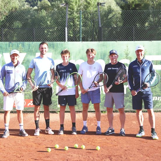 Sechs Männer stehen auf einem Tennisplatz, halten Tennisschläger in der Hand, mit Tennisbällen auf dem Boden verstreut. Sie sind in Freizeitkleidung gekleidet und scheinen für ein Foto zu posieren.