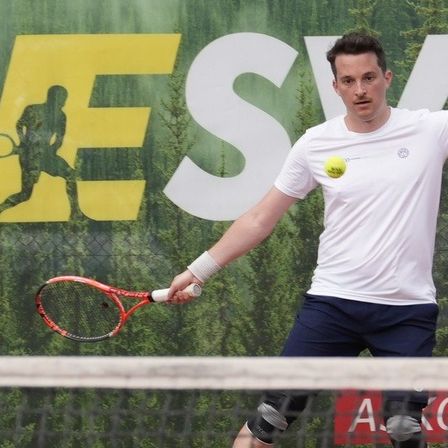 Ein Mann in einem weißen T-Shirt spielt Tennis auf einem Platz mit grünem Hintergrund, der eine Tennisclub-Werbung zeigt.