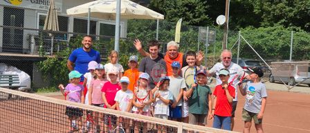 Eine Gruppe von Menschen, darunter Kinder, steht auf einem Tennisplatz. Einige halten Tennisschläger. Dahinter sind ein Zaun, ein Gebäude und Bäume zu sehen.