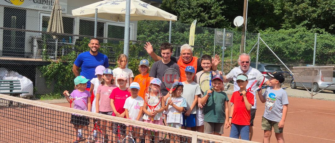 Eine Gruppe von Menschen, darunter Kinder, steht auf einem Tennisplatz. Einige halten Tennisschläger. Dahinter sind ein Zaun, ein Gebäude und Bäume zu sehen.