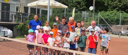 Eine Gruppe von Kindern und Erwachsenen posiert für ein Foto auf einem Tennisplatz mit einem Netz und einem Gebäude dahinter.