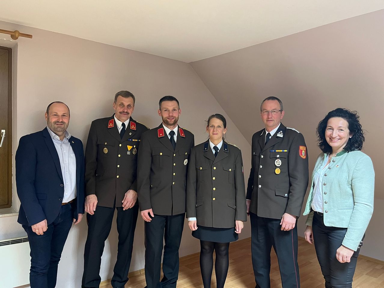 Fünf Personen, in Uniformen gekleidet, stehen in einem Raum zusammen. Drei Männer und zwei Frauen, alle lächeln. Der Mann ganz links trägt einen Anzug. Die anderen tragen Uniformen mit Abzeichen.