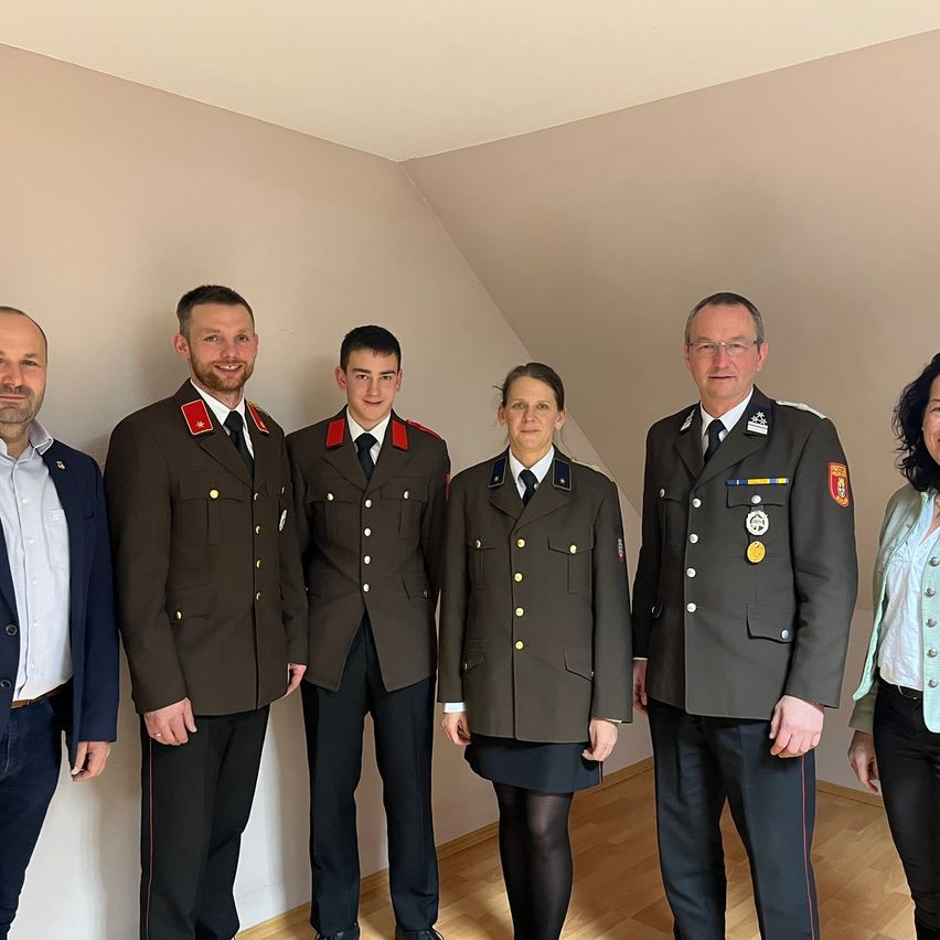 Sechs Personen in Uniformen posieren für ein Foto in einem Raum. Drei Männer und eine Frau tragen Militäruniformen, während die anderen beiden Männer formelle Kleidung tragen.