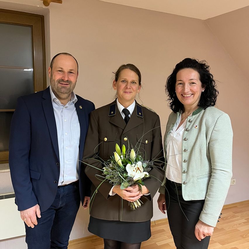 Drei Personen stehen eng beieinander und lächeln für ein Foto. Die Frau in der Mitte hält einen Blumenstrauß. Der Mann auf der linken Seite trägt einen Anzug und die Frau auf der rechten Seite trägt eine grüne Jacke.