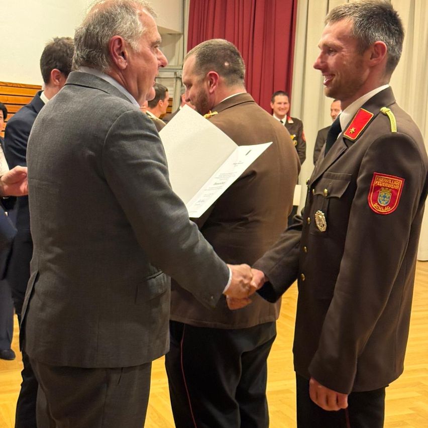 Two men in uniforms are shaking hands in an indoor setting. One man holds a piece of paper. A few people stand behind them.