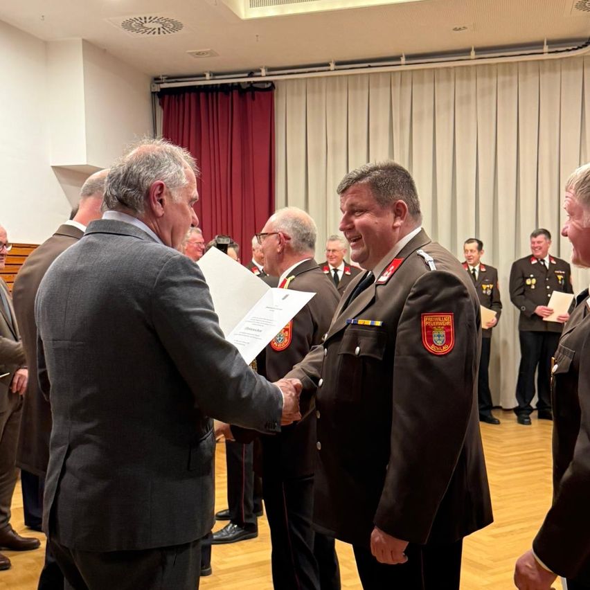 A group of people dressed in formal attire are gathered in a hall. One man shakes hands with another. Some people are standing behind them. A few people are holding papers.