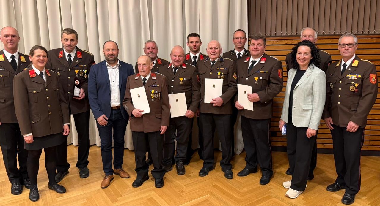 A group of men, some in military uniforms, stand together holding certificates. They are smiling and posing for a photo. Behind them is a curtain, and some are wearing hats.