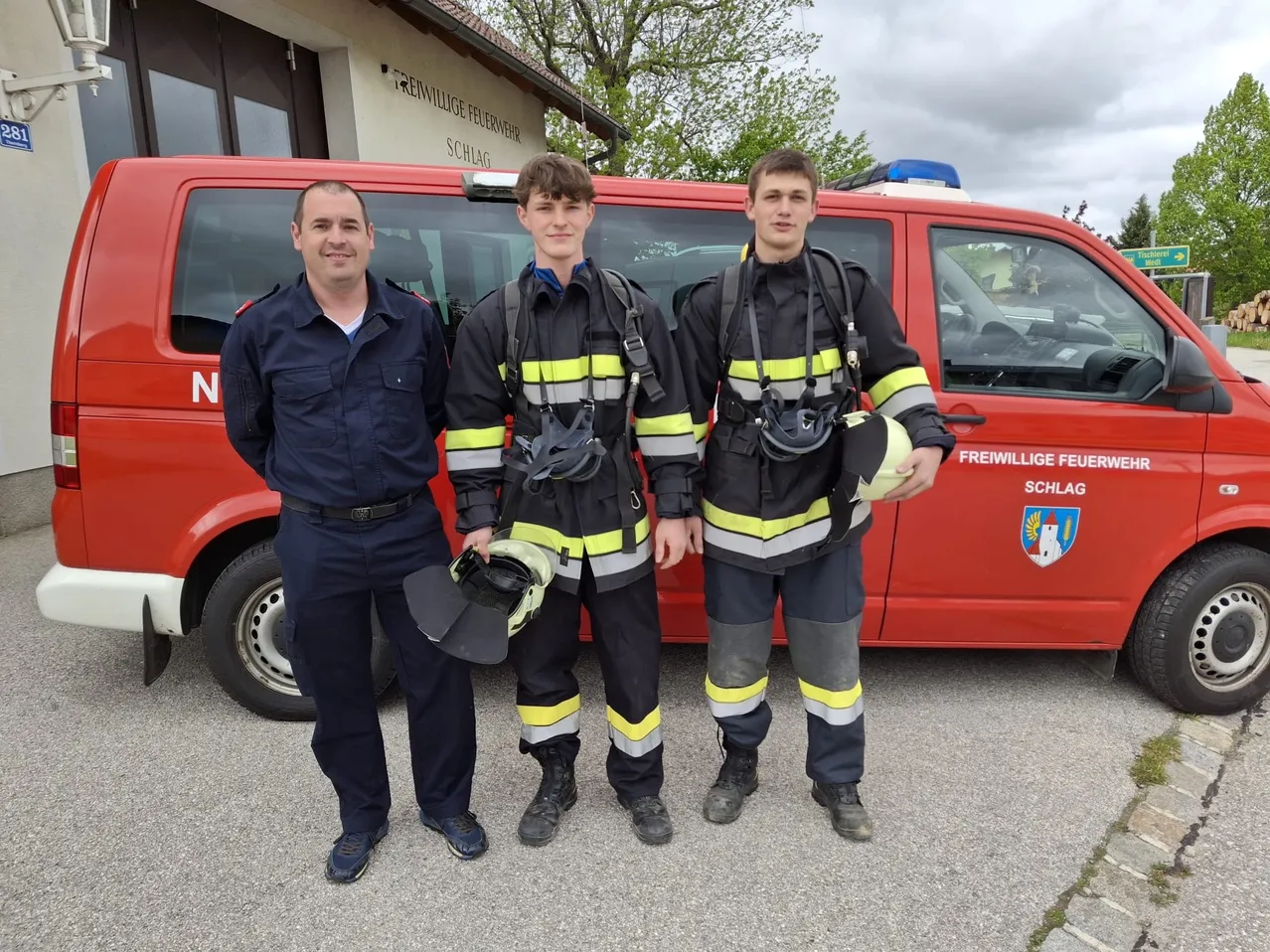Drei Männer, zwei in Feuerwehruniformen und einer in einer dunkelblauen Uniform, posieren für ein Foto vor einem roten Feuerwehrwagen.