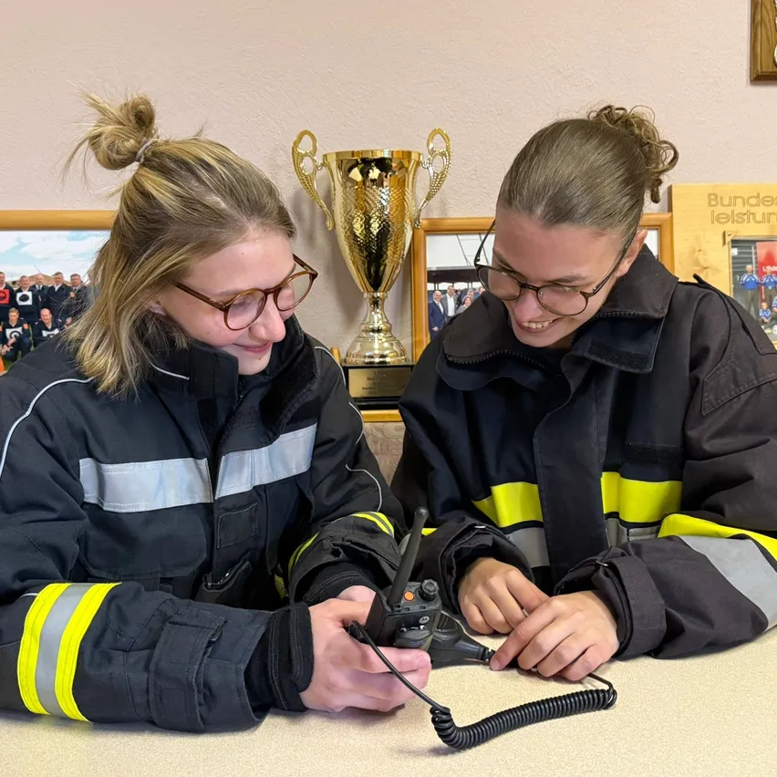Zwei Frauen in Feuerwehrjacken sitzen an einem Tisch, eine bedient ein Funkgerät. Sie sind umgeben von gerahmten Fotos und einem Pokal.