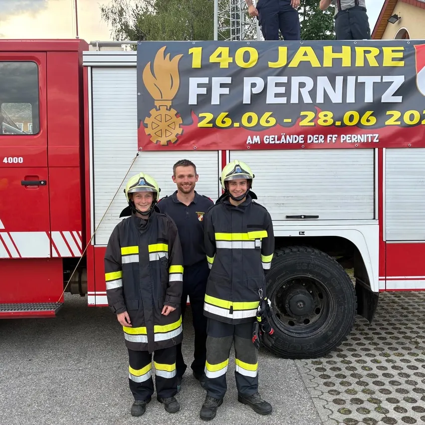 Drei Feuerwehrleute, darunter ein lächelndes Kind, stehen vor einem roten Feuerwehrauto mit einem Schild, das 140 Jahre der Feuerwehr Pernitz feiert.