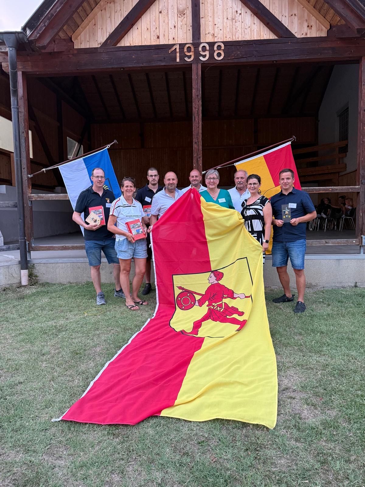 Eine Gruppe von Menschen steht vor einem Gebäude mit Fahnen, hält eine große Fahne und Bücher und lächelt für ein Foto.