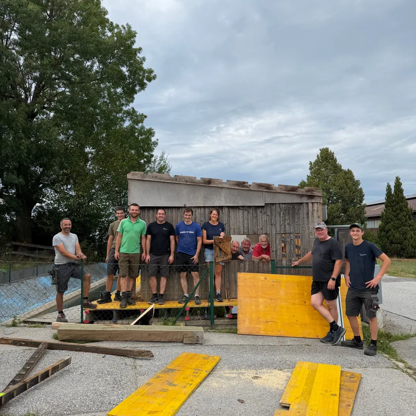 Eine Gruppe von Menschen, sowohl Männer als auch Frauen, stehen auf einer Holzplattform vor einem kleinen Holzgebäude. Sie sind alle in Freizeitkleidung gekleidet. Einige halten sich an der Umzäunung fest, während andere auf der Plattform stehen. Es gibt Holzbretter auf dem Boden und einen Baum im Hintergrund.