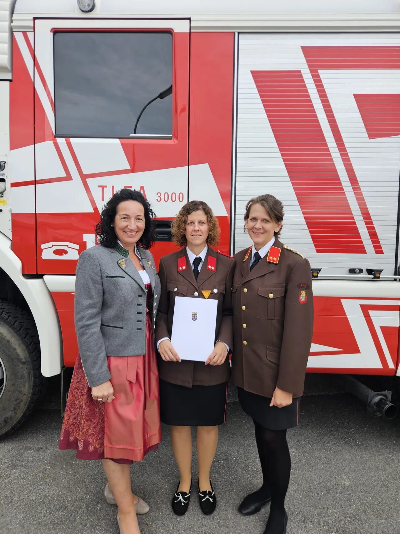 Drei Frauen stehen vor einem Feuerwehrauto, eine hält ein Zertifikat. Die Frau links trägt einen grauen Blazer und einen roten Rock. Die Frau in der Mitte hält ein Zertifikat und die Frau rechts trägt eine braune Uniform.