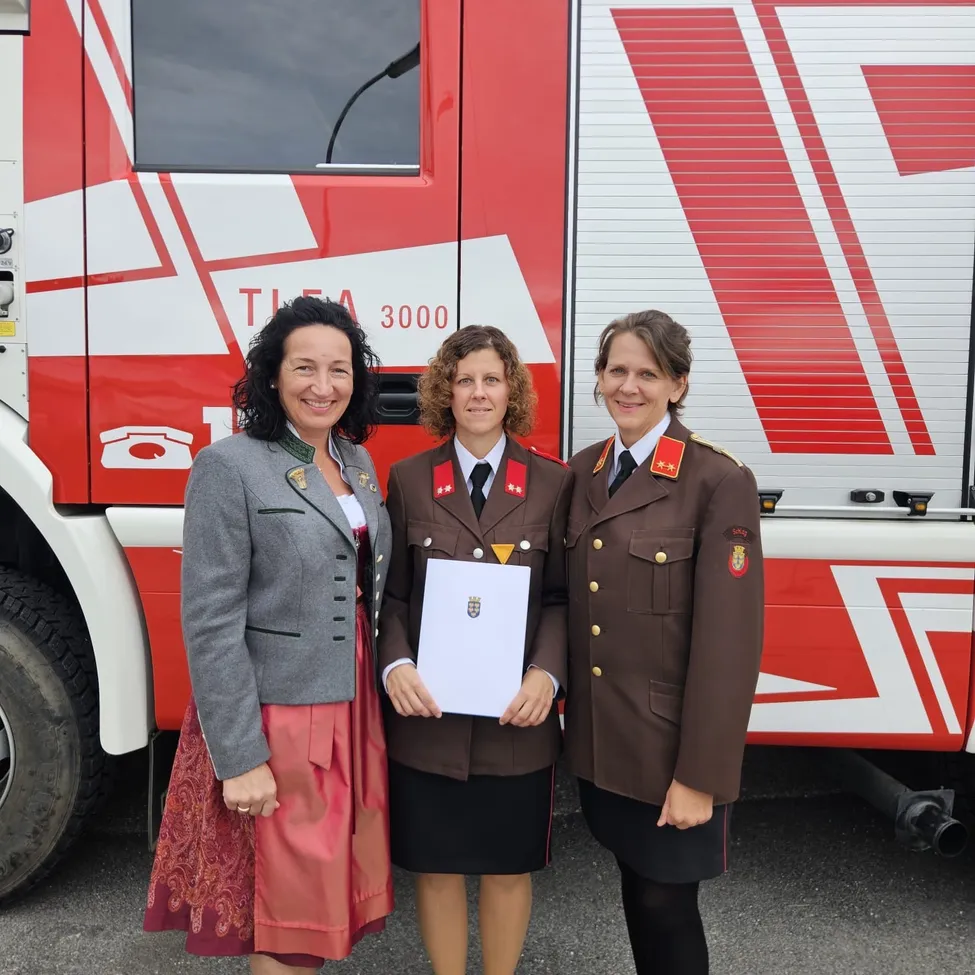 Drei Frauen stehen vor einem Feuerwehrauto, eine hält ein Zertifikat. Die Frau links trägt einen grauen Blazer und einen roten Rock. Die Frau in der Mitte hält ein Zertifikat und die Frau rechts trägt eine braune Uniform.