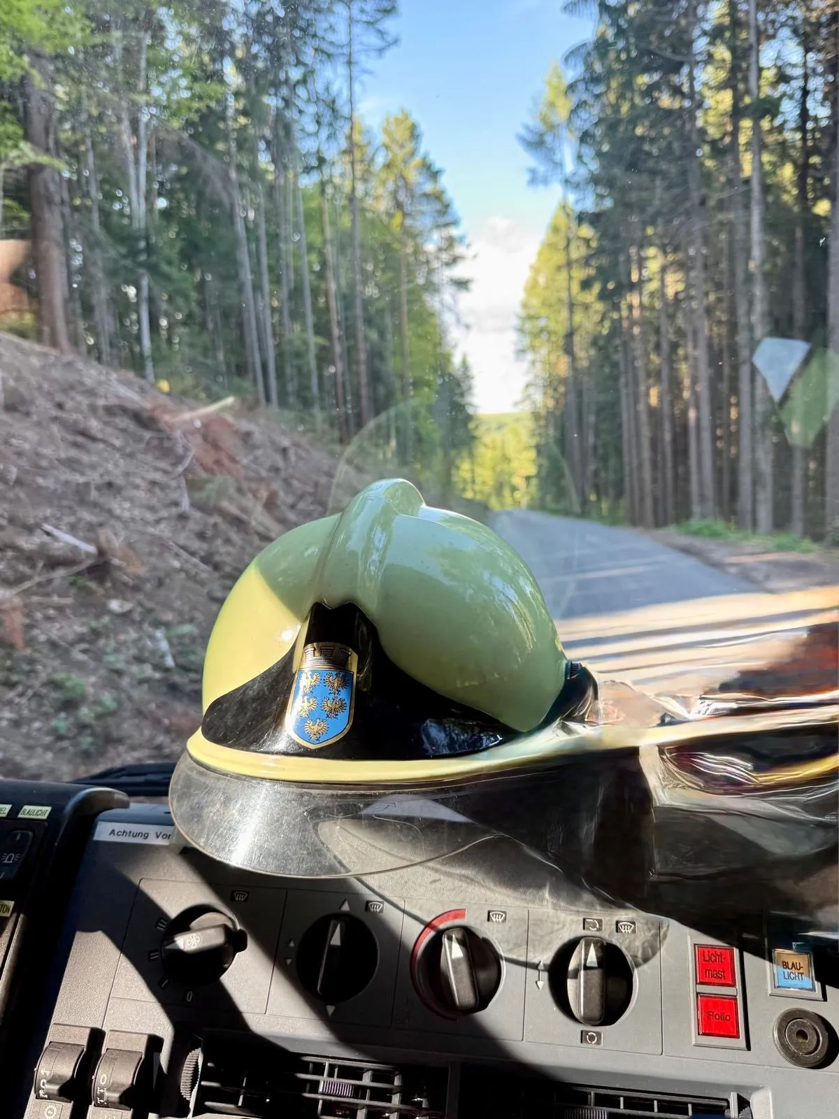 Ein grüner Helm mit blauem Emblem ist auf einem Armaturenbrett zu sehen, mit Blick auf eine Straße und einen Wald im Hintergrund.