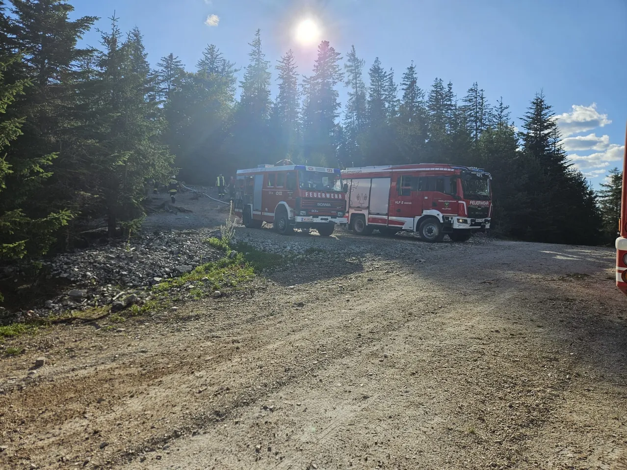 Zwei rote Feuerwehrfahrzeuge sind auf einer unbefestigten Straße zwischen Bäumen geparkt. Eines ist mit 'FEUERWEHR' beschriftet, und eine Person ist in der Nähe. Die Sonne scheint hell.