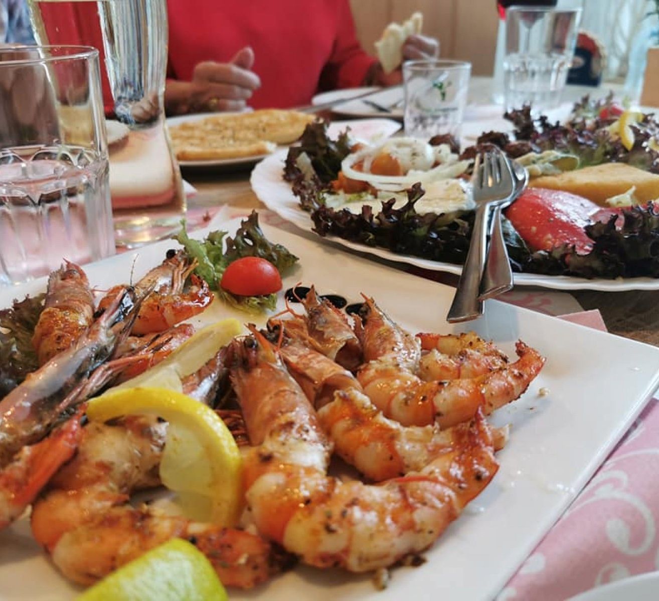 Ein Tisch mit verschiedenen Gerichten, darunter gegrillte Garnelen, Salat und Brot. Eine Person sitzt im Hintergrund. Es gibt Gläser mit Wasser und Wein auf dem Tisch.