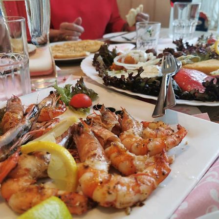 Ein Tisch mit verschiedenen Gerichten, darunter gegrillte Garnelen, Salat und Brot. Eine Person sitzt im Hintergrund. Es gibt Gläser mit Wasser und Wein auf dem Tisch.