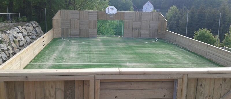 Ein erhöhtes Basketballfeld im Freien mit Holzzäunen, einem grünen Feld und einem Haus in der Ferne.