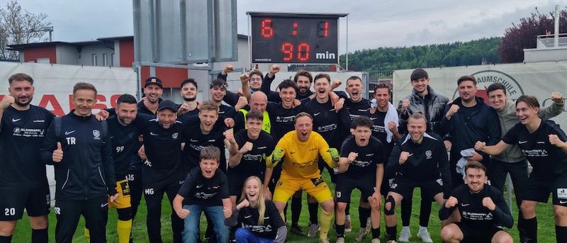 Ein Fußballteam feiert einen Sieg auf einem Feld, wobei die Anzeigetafel 6:1 zeigt und das Endergebnis 6-1 lautet. Die Teammitglieder tragen schwarze Trikots und einige von ihnen gelbe Trikots.