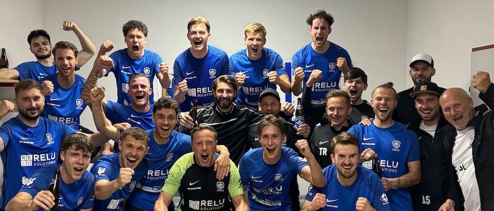 Eine Fußballmannschaft in blauen Trikots, darunter ein Spieler in einem schwarz-gelben Trikot, feiert zusammen in einer Umkleidekabine, einige halten Getränke.