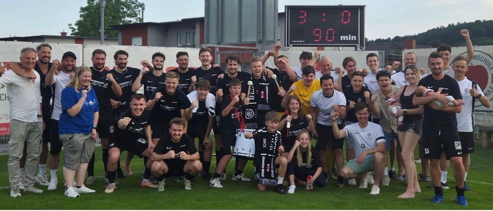 Ein Sportteam feiert einen Sieg auf einem Feld, wobei die Anzeigetafel 3:0 zeigt. Sie halten Trophäen und Trommeln, einige tragen Trikots. Dahinter befinden sich Gebäude und Bäume.