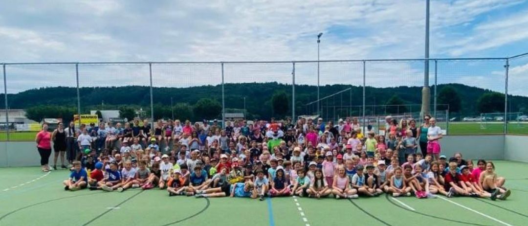 Ein Sportfest an der Volksschule St. Ruprecht/R. Eine große Gruppe von Kindern und Erwachsenen versammelt sich auf einem Sportfeld mit Zaun. Viele Vereine unterstützten die Veranstaltung.