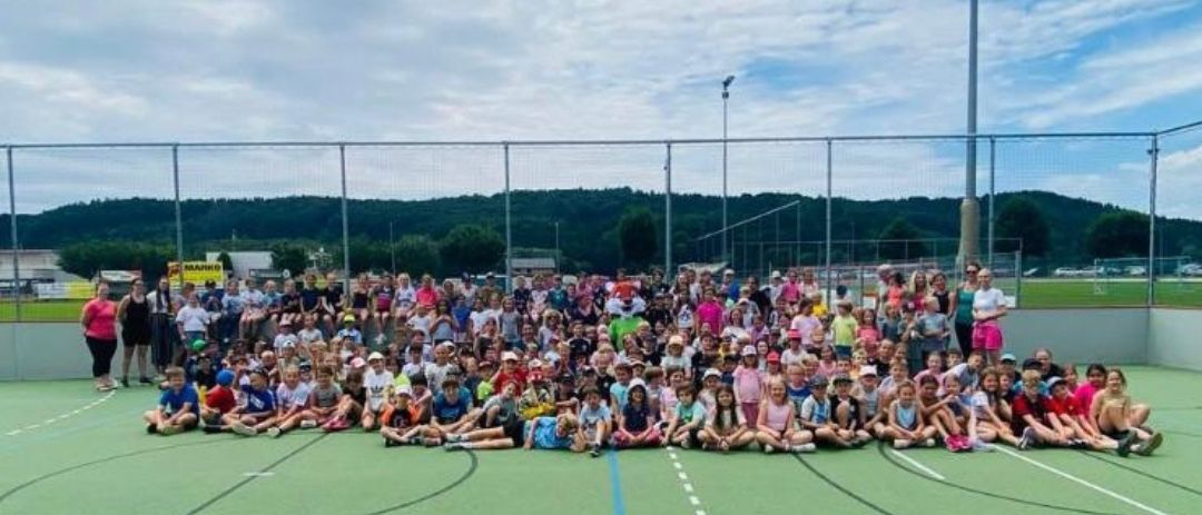 Ein Sportfest an der Volksschule St. Ruprecht/R. Eine große Gruppe von Kindern und Erwachsenen versammelt sich auf einem Sportfeld mit Zaun. Viele Vereine unterstützten die Veranstaltung.