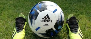 Ein Adidas-Fußball mit der Aufschrift Team Junior 350 liegt zwischen zwei neongelben Adidas-Fußballschuhen auf einem Rasenfeld.