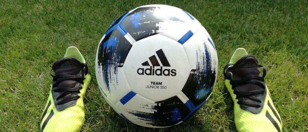 Ein Adidas-Fußball mit der Aufschrift Team Junior 350 liegt zwischen zwei neongelben Adidas-Fußballschuhen auf einem Rasenfeld.