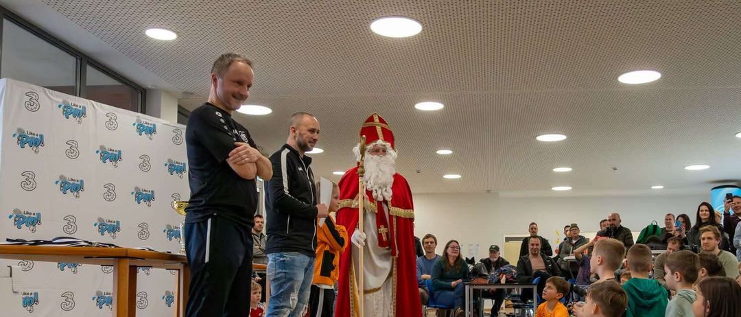 Ein Mann in Santa Claus-Kostüm steht mit zwei Männern in einer Sporthalle. Sie sind von einer Menge Kinder und Erwachsener umgeben, einige sitzend und andere stehend.