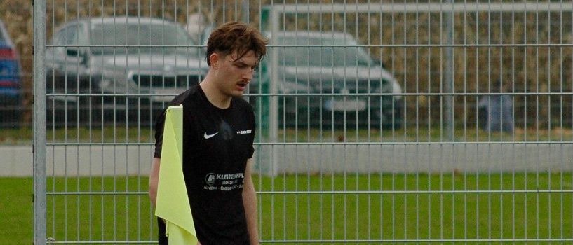 Ein Fußballspieler in schwarzer Uniform steht neben einem Torpfosten mit einem Fußball auf dem Boden.