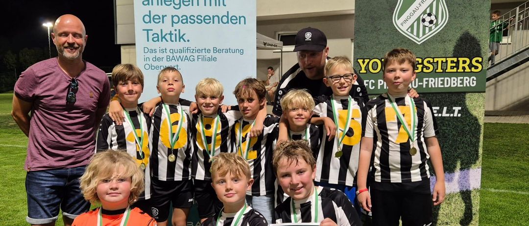 Eine Gruppe junger Jungen in Sportuniformen mit Medaillen um den Hals posiert für ein Foto auf einem Rasenfeld. Sie lächeln und schauen in die Kamera. Hinter ihnen steht ein Mann mit Hut und Brille. Auf der linken Seite befindet sich ein Banner mit Text und Logo.
