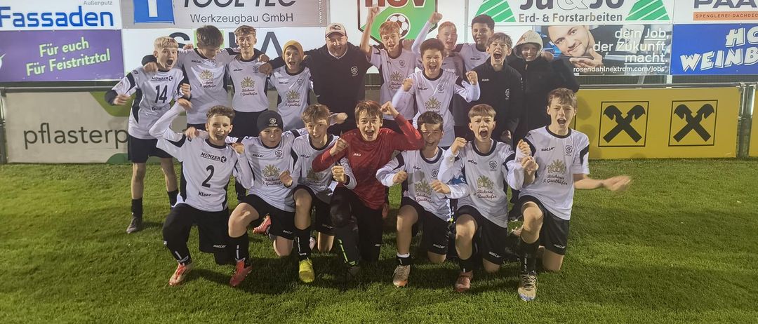 Eine Fußballmannschaft feiert auf einem Feld mit einem Anzeigetafel im Hintergrund, die das Endergebnis zeigt. Sie tragen alle weiße Trikots mit Nummern. Im Hintergrund befinden sich Werbebanner.