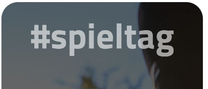 Ein Fußball mit roten und weißen Flächen steht auf einem Rasenfeld, mit einem verschwommenen Hintergrund von Bäumen und einem verschwommenen Bein eines Menschen im Vordergrund.
