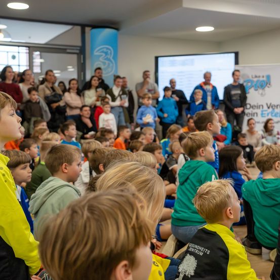 In einem großen Raum versammeln sich Kinder und Erwachsene, um eine Präsentation zu beobachten. Die Kinder sitzen vorne, einige tragen Jacken mit Logos, während andere dahinter stehen. Eine Projektionsleinwand zeigt Informationen, und ein Banner mit dem Text 'Like a Pro' ist sichtbar.