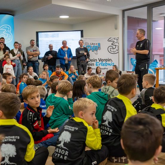 Ein Mann steht vor einer Gruppe von Kindern in einem Raum, wahrscheinlich einem Auditorium. Die Kinder sitzen auf dem Boden und hören aufmerksam zu. Hinter dem Mann befindet sich eine Projektionsleinwand und ein Banner. Einige Erwachsene stehen im Raum herum.